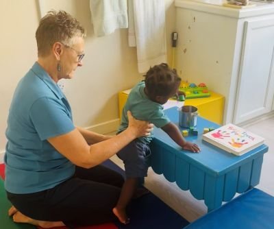 Image of physiotherapist and child. The therapist is sitting on the floor behind the child supporting his back as he stands leaning against a short table.