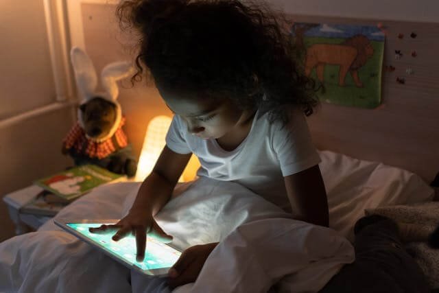 Image of a young black child sitting up in bed at night, while playing a game on her tablet.