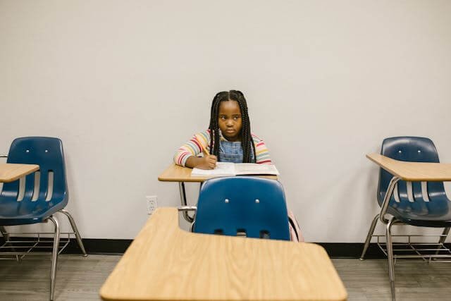 Image of a young black girl sitting at a school desk writing in a noteboook.