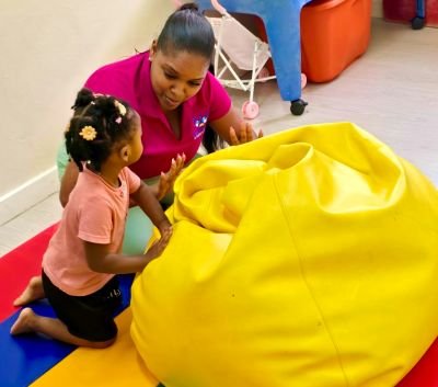 Image of the occupational therapist and a child. They are on a play mat pushing a large bean bag together and making eye contact.