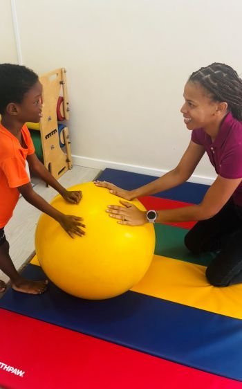 Image of the Floortime™ practitioner and a child. They are on a play mat with both of their hands on a therapy ball between them while making eye contact and smiling.